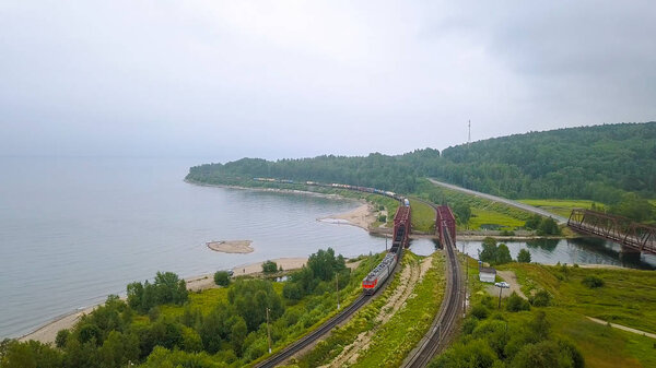 Транссибирская железная дорога, побережье Байкала. Движение поездов по железному мосту через реку, впадающую в Байкал. Россия, с дрона
  