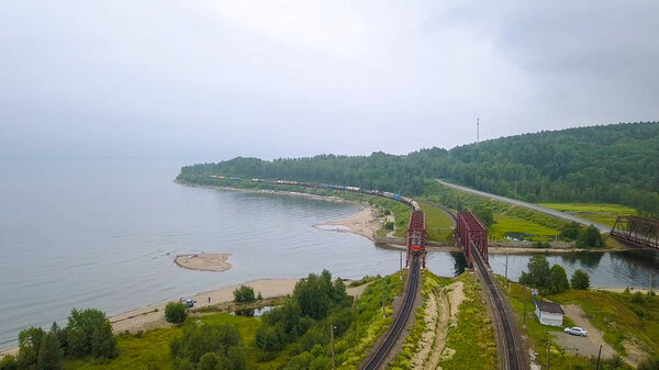 Транссибирская железная дорога, побережье Байкала. Движение поездов по железному мосту через реку, впадающую в Байкал. Россия, с дрона
 