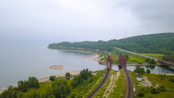 Транссибирская железная дорога, побережье Байкала. Движение поездов по железному мосту через реку, впадающую в Байкал. Россия, с дрона
 