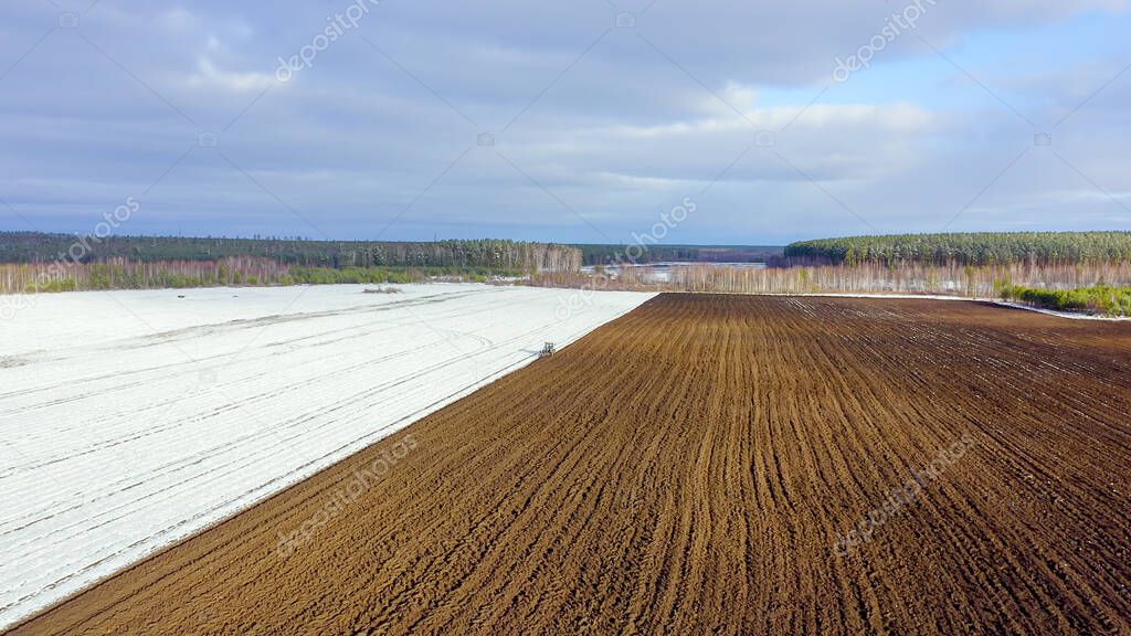 A blue tractor plows a field covered with snow. Behind the tractor is black earth. Russia, Ural, Aerial View  