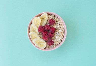 Düz bir güzel pembe kase yulaf ezmesi ile taze tatlı rasberry ve muz dilimleri turkuaz arka plan üzerinde göklere.