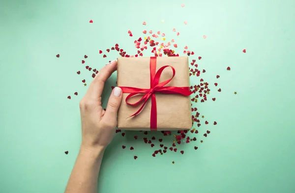 Stock image Gift box wrapped in brown colored craft paper and tied with red bow on blue background with red confetti. Lady's hand holding a giftbox.