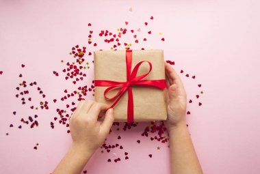 Çocuk kahverengi renkli Kraft kağıt sarılı ve kırmızı kalp şekli konfeti ile pembe arka plan üzerinde kırmızı yay ile bağlı hediye kutusu tutuyor.