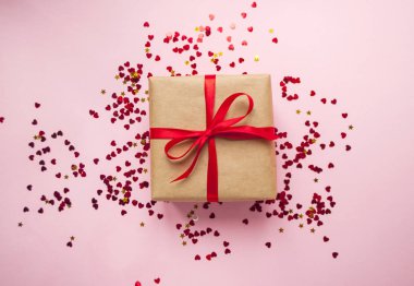 Hediye kutusu kahverengi renkli Kraft kağıt sarılı ve kırmızı konfeti ile pastel pembe arka plan üzerinde kırmızı yay ile bağlı.