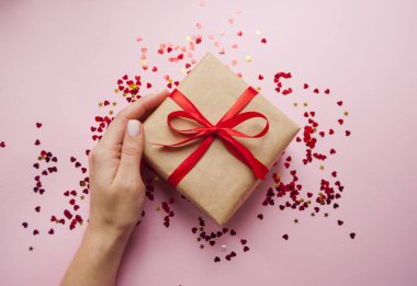 Hediye kutusu kahverengi renkli Kraft kağıt sarılı ve kırmızı konfeti ile pembe arka plan üzerinde kırmızı yay ile bağlı. Bayanın elindeki bir giftbox holding.