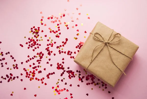 Kahverengi renkli Kraft kağıt sarılı ve kalp şekli kırmızı konfeti ile pembe arka plan üzerinde iple bağlı hediye kutusu.
