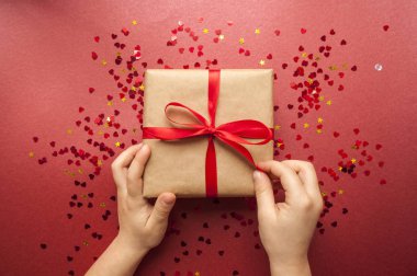 Çocuk kahverengi renkli Kraft kağıt sarılı ve kırmızı kalp şekli konfeti koyu kırmızı zemin üzerine kırmızı yay ile bağlı hediye kutusu tutuyor.