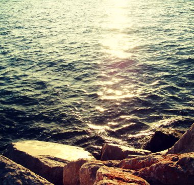 Yeşil deniz yüzeyi ve kayalık kıyı, yumuşak odak yansıtan günbatımı güneş ışınları ile arka ışık