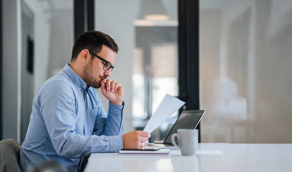 Serious pensive thoughtful young businessman or entrepreneur in modern contemporary office looking at and working with laptop and paper