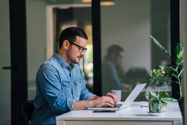 Serious pensive thoughtful focused young casual businessman or entrepreneur in office looking at and working with laptop making and typing serious important business email — Stock Photo, Image