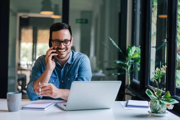 Retrato de un joven empresario alegre sonriente en una oficina informal haciendo una llamada telefónica mientras trabaja con una computadora portátil — Foto de Stock