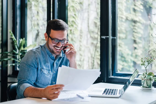 Портрет молодого улыбающегося жизнерадостного предпринимателя в офисе, делающего телефонный звонок во время работы с графиками и графиками, глядя на бумажные документы — стоковое фото