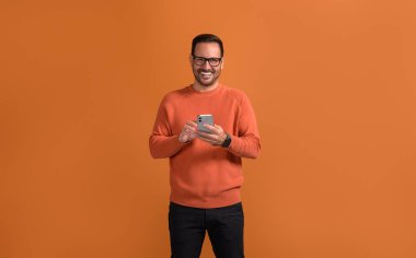 Akıllı telefondan gülümseyen yakışıklı profesyonel mesajlaşma turuncu arka planda güvenle poz verirken.