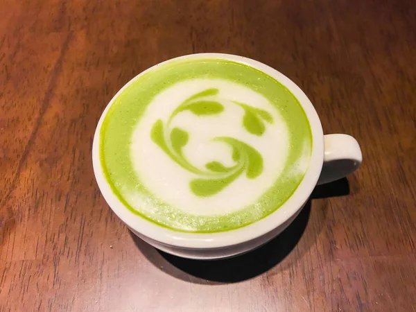 Hot matcha green tea milk latte with creamy milk is flower or plant pattern, a little sugar and teaspoon in a cup on the wooden table.