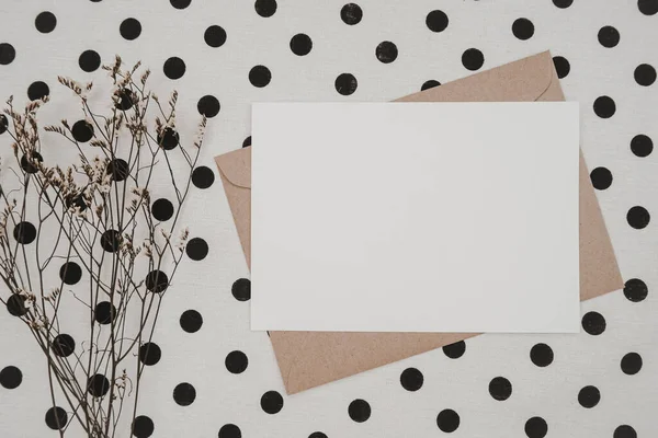 Blank white paper on brown paper envelope with Limonium dry flower and Carton box on White cloth with black dots. Mock-up of horizontal blank greeting card. Top view of Craft  envelope. Flat lay.