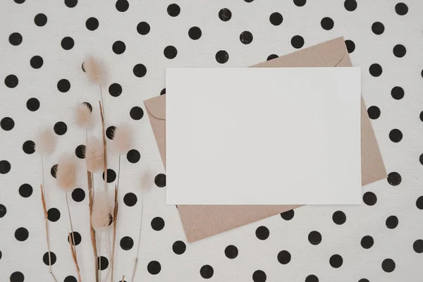 Blank white paper on brown paper envelope with Rabbit tail dry flower and Carton box on White cloth with black dots. Mock-up of horizontal blank greeting card. Top view of Craft  envelope. Flat lay.