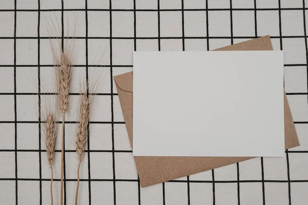 Blank white paper on brown paper envelope with Barley dry flower and Carton box on White cloth with Black grid pattern. Mock-up of horizontal blank greeting card. Top view of Craft envelope. Flat lay