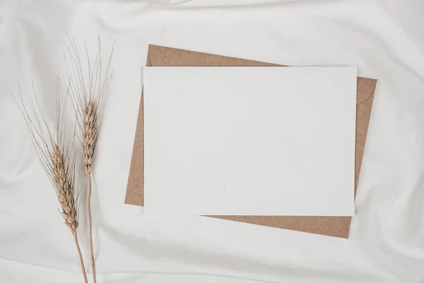 Blank white paper on brown paper envelope with Barley dry flower on white cloth. Mock-up of horizontal blank greeting card. Top view of Craft envelope on white background. Flat lay minimalism
