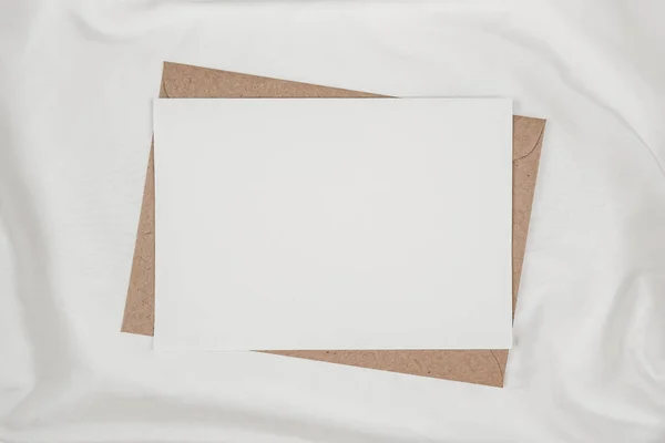 Blank white paper on brown paper envelope on white cloth. Mock-up of horizontal blank greeting card. Top view of Craft envelope on white background. Flat lay minimalism