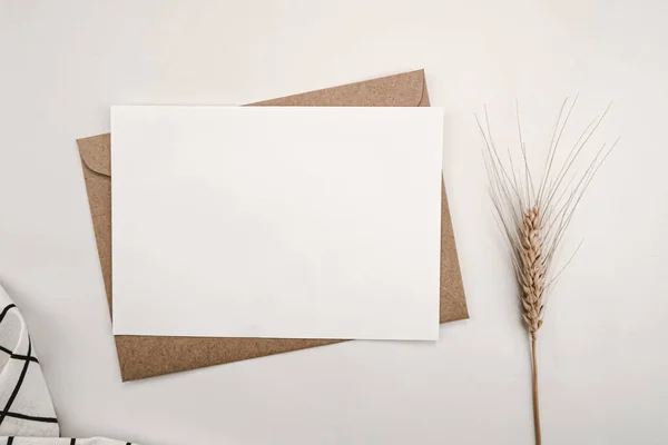 Blank white paper on brown paper envelope with Barley dry flower and White cloth with Black grid pattern. Mock-up of horizontal blank greeting card. Top view of Craft envelope on white background.