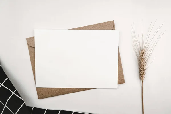 Blank white paper on brown paper envelope with Barley dry flower and and Black cloth with White grid pattern. Mock-up of horizontal blank greeting card. Top view of Craft envelope on white background.