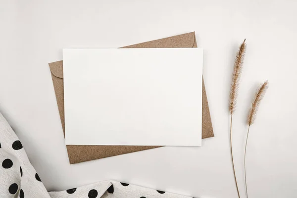 Blank white paper on brown paper envelope with Bristly foxtail dry flower and White cloth with black dots. Mock-up of horizontal blank greeting card. Top view of Craft envelope on white background.