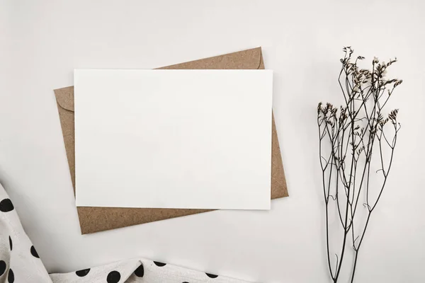 Blank white paper on brown paper envelope with Limonium dry flower and White cloth with black dots. Mock-up of horizontal blank greeting card. Top view of Craft envelope on white background.