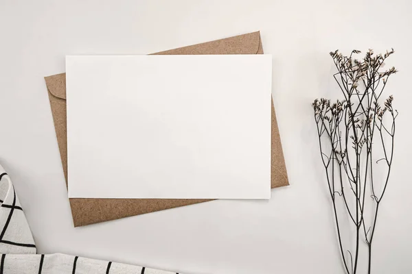 Blank white paper on brown paper envelope with Limonium dry flower and White cloth with Black grid pattern. Mock-up of horizontal blank greeting card. Top view of Craft envelope on white background.