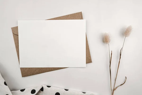 Blank white paper on brown paper envelope with Rabbit tail dry flower and White cloth with black dots. Mock-up of horizontal blank greeting card. Top view of Craft envelope on white background.