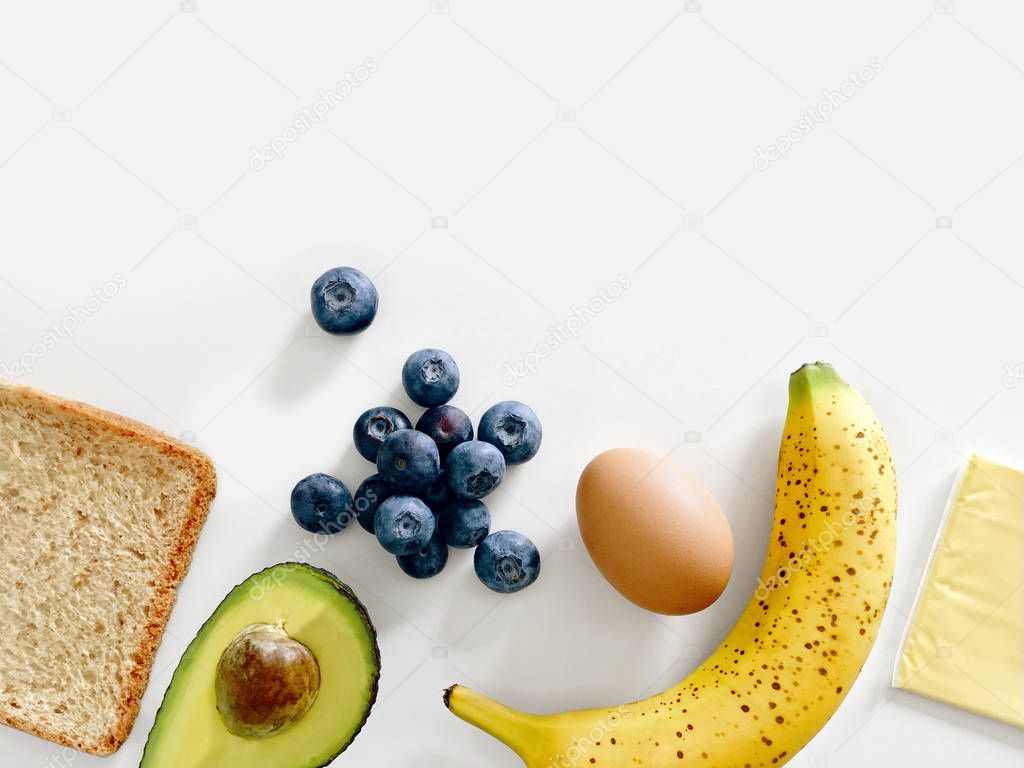 Healthy breakfast ingredients with whole wheat bread, avocado, blueberries, banana, egg, and cheese isolated on a white background, top view with copy space