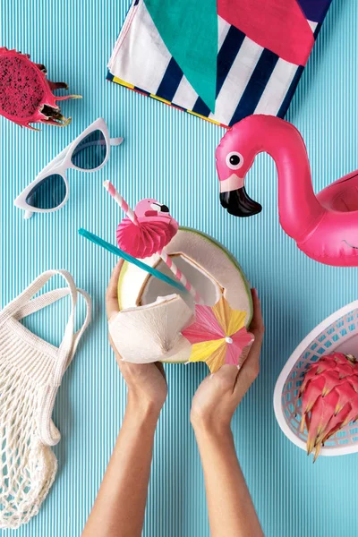 Flat lay of summer vibes concept with colorful travel items, sunglasses, scarf, coconut water, pink dragon fruit, flamingo inflatable drink holder and net bag on a blue background.