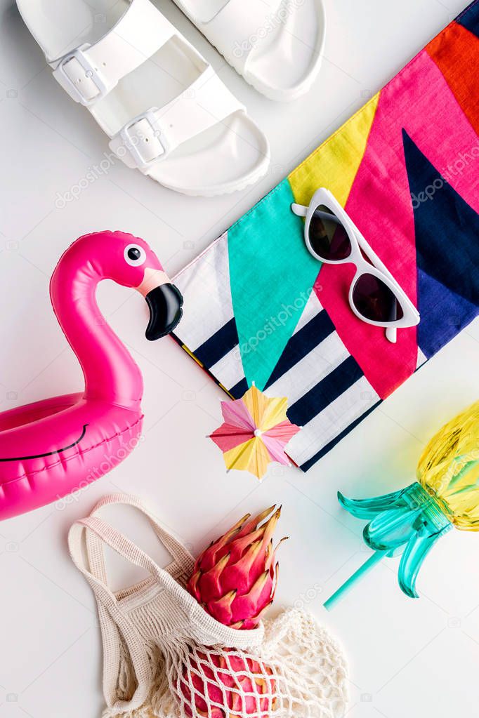 Flat lay of summer vibes concept with colorful travel fashion items, sunglasses, scarf, pink dragon fruit, flamingo inflatable drink holder, pineapple straw tumbler bottle, net bag and sandal on a white background.