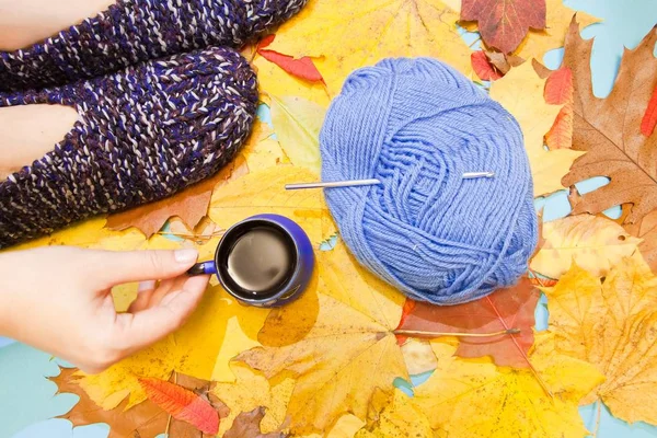 Cool  flat lay for creative people who love hand made and knitting - warm comfortable knitted slippers, a hand holding a cup of hot coffee, and a ball of woolen or cotton yarn with crochet hook. Autumn yellow and orange leaves background, studio phot