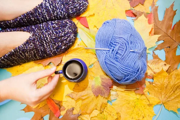 Cool  flat lay for creative people who love hand made and knitting - warm comfortable knitted slippers, a hand holding a cup of hot coffee, and a ball of woolen or cotton yarn with crochet hook. Autumn yellow and orange leaves background, studio phot