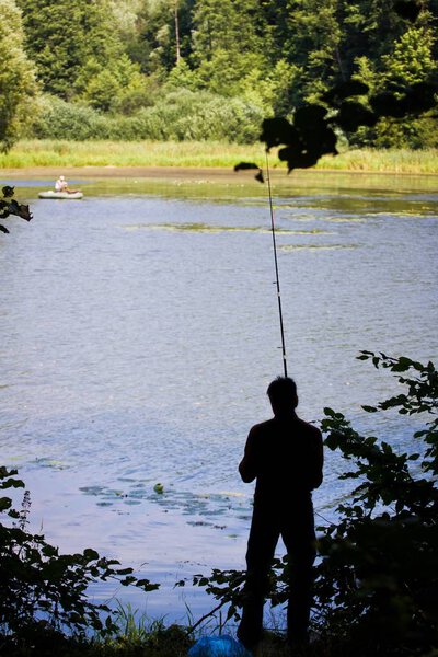 fisherman's figure with  a fishing rod on a bank of little natural lake in forest, calm breeze on water surface, hobby leisure time image