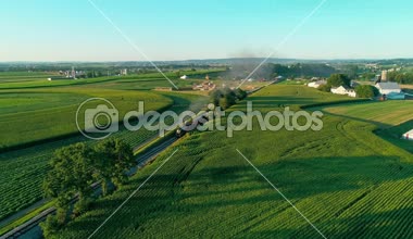 Tren geçerken Amish çiftlik arazileri ve kırsal yoluyla bir geç günbatımı yaz gününde dron tarafından görüldüğü gibi Buhar