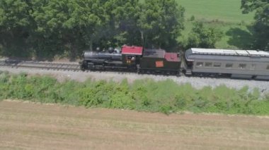 Ronks, Pennsylvania, Temmuz 2019 - Bir Drone tarafından görüldü olarak Güneşli bir Yaz Günü Amish Kırsal Boyunca Seyahat Yolcu Tren Puffing Duman ile 1910 Buhar Motoru Havadan Görünümü