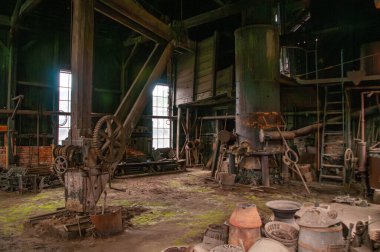 Eski bir endüstriyel demir ocağı atölyesinde paslı makineler dağınık aletlerin arasında duruyordu. Kirli pencerelerden süzülen güneş ışığı tozlu zeminde yeşilimsi bir renk bırakıyor ve nostaljik bir hava yaratıyor.