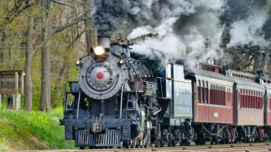 Tarihi bir buhar lokomotifi, yemyeşil ağaçlarla çevrili raylar boyunca klasik yolcu vagonlarını çeker. Kalın siyah duman taşraya doğru hareket ederken motordan yükseliyor..