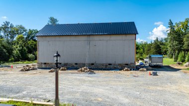 Kırsal bölgedeki büyük bir ahırda çalışmalar sürüyor. Malzemeler yakınlarda düzenlenirken bir merdiven binaya dayanıyor. Gökyüzü açık ve mavidir, huzurlu ortamı güçlendirir..