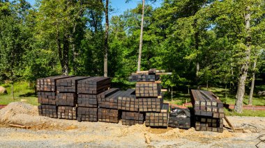 Yeşil ağaçlarla çevrili bir inşaat alanında büyük bir kereste yığını düzgünce düzenlenir. Parlak güneş ışığı, sağlam ahşap kirişleri vurguluyor. Bu da inşaat projelerine hazırlık olduğunu gösteriyor..