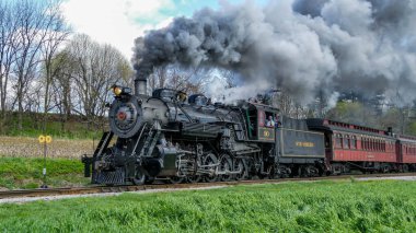 Ronks, Pennsylvania, 4-13-2025 - Güçlü bir buhar lokomotifi yemyeşil alanlarda seyahat ederek açık mavi gökyüzüne duman bulutları salıyor. Yolcular bu yolculukta manzaranın tadını çıkarıyorlar.