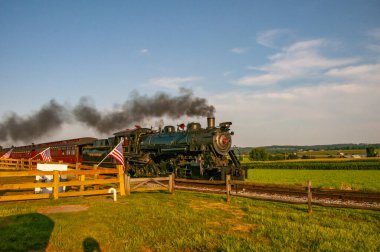 Ronks, Pennsylvania, 3 Ağustos 2025 öğleden sonra yeşil tarlalardan geçen klasik buhar treni. Lokomotif, ahşap çitlerin önünden geçerken siyah duman püskürtür..