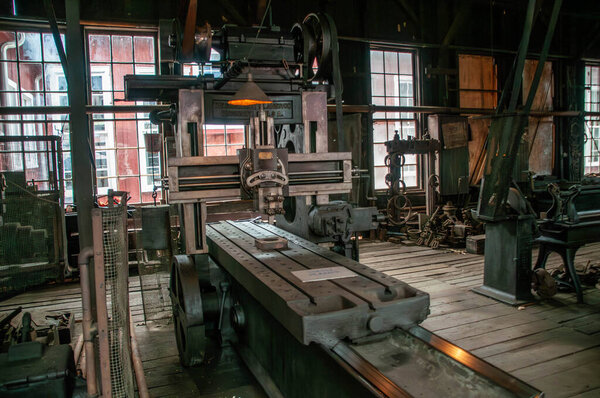 A large vintage milling machine sits prominently in an industrial workshop, surrounded by various tools and equipment. Sunlight streams through the windows, illuminating the workspace.