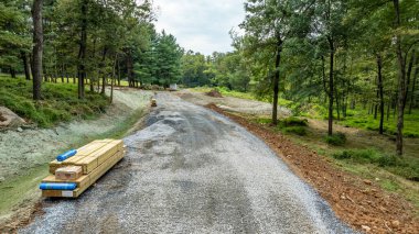 İşçiler ağaçlar ve yeşilliklerle çevrili yeni bir yolu tamamlıyorlar. Kereste ve inşaat malzemeleri düzgün bir şekilde yakınlarda istiflenmiş. Bu da doğal ortamdaki gelişimin devam ettiğini gösteriyor..