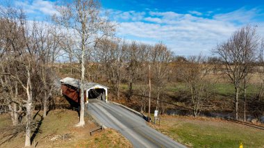 Etrafı yapraksız ağaçlarla çevrili dolambaçlı bir yolda kırsal bir köprü manzarası. Parlak mavi gökyüzü, ilkbaharın başlarındaki topraksı tonlara katıksız bir tezat sağlar..