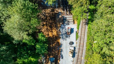 Heavy machinery is at work along railway tracks surrounded by dense trees. The site shows preparation for infrastructure development under clear midday skies.