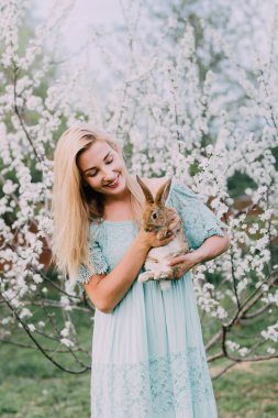Beyaz bahar bahçesinde bir tavşan tutan bir mavi elbise gülüyor kız.