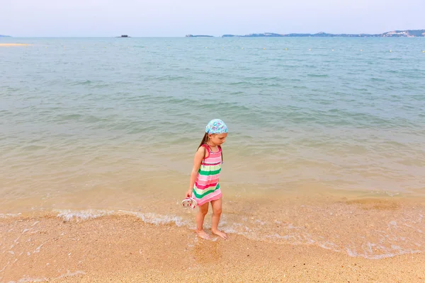 Ένα κοριτσάκι με ριγέ φόρεμα περπατά κατά μήκος της παραλίας σε ένα ηλιόλουστο απόγευμα. — Φωτογραφία Αρχείου