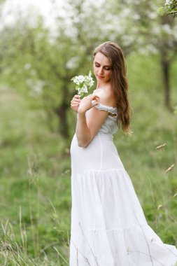 Bahçedeki hamile gelin. Beyaz boho tarzı gelinlik. Yeşil ağaçlar ve güneş. Beyaz elbiseli güzel hamile kız bahar öğleden sonra çiçekli bir bahar parkta yürüyor.